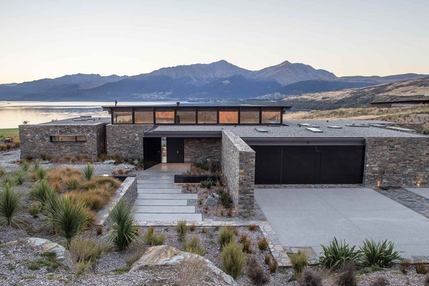 A low, modern single-storey residence set in a rugged landscaped site overlooking a lake and mountain range. The building features dark flat-roof volumes, extensive upper clerestory glazing, and walls clad in irregular grey stone. A broad concrete driveway leads to a double garage, while a stepped paved entry path passes through drought-tolerant grasses and gravel landscaping.