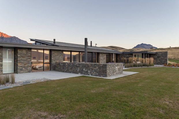 Low, single-storey contemporary residence set in an open mountainous landscape. The building has extensive glazing, dark stone masonry walls, a shallow low-pitched roof, and a central enclosed courtyard or terrace with a stone privacy wall. Native grasses and restrained landscaping reinforce the connection to the surrounding alpine setting.