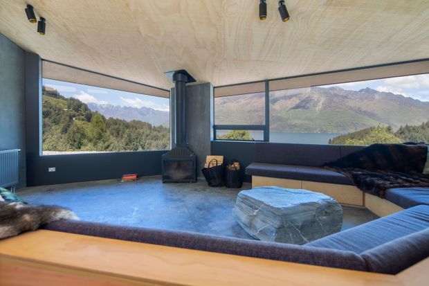 A contemporary living room with expansive corner glazing overlooking a mountain and lake landscape. The room features a low built-in sectional sofa, a sculptural stone coffee table, a black freestanding wood-burning stove, dark window frames, and a plywood-lined ceiling with recessed spotlights. A dark concrete-like floor and natural materials create a warm, rugged retreat atmosphere.