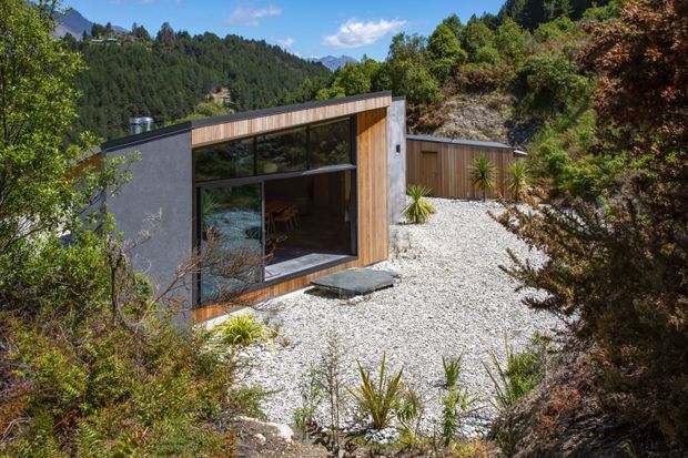 A compact contemporary cabin set into a steep, bush-covered mountain site. The single-storey building has dark painted wall cladding, warm vertical timber cladding, a low mono-pitched roof, and expansive glazed openings. A pale gravel landscape with native-style plantings surrounds the cabin, with forested hills and distant mountains visible beyond.