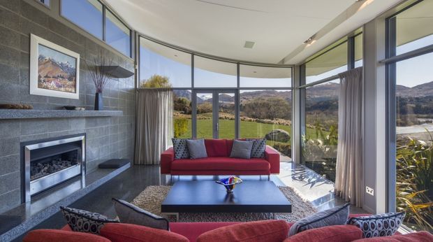 A contemporary living room with expansive curved floor-to-ceiling glazing overlooking a green valley and hills. The room features a red sofa and armchairs, a large dark coffee table, a grey rug, a substantial grey tiled fireplace wall, and neutral curtains. The curved ceiling and panoramic windows create a bright, open connection to the landscape.