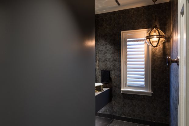 A moody, compact bathroom or powder room interior viewed partly through a doorway. The room has dark textured wall finishes, dark tiled flooring, a white-framed window with plantation shutters, and a spherical pendant light with a metal cage. A partial vanity or basin is visible at left.
