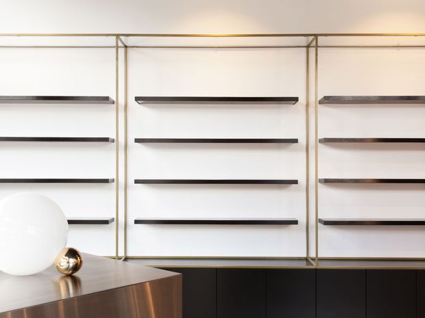 A refined contemporary built-in display and storage wall with evenly spaced dark floating shelves framed by slim brass-toned metal trim. A dark lower cabinetry run sits beneath the shelving, while a sculptural white globe lamp and metallic console occupy the foreground. The composition is minimal, polished, and high contrast.