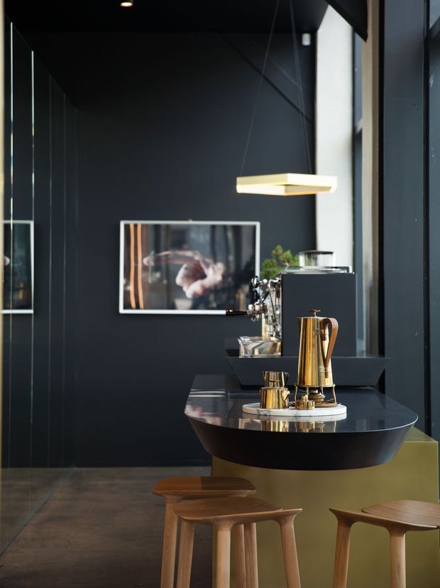 A sophisticated dark-toned kitchen or home bar interior with a sculptural black island and polished dark benchtop, brass coffee equipment, timber stools, integrated appliances, black walls, reflective cabinetry, and a contemporary rectangular pendant light. A framed artwork and indoor greenery add visual interest.