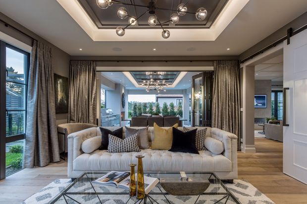 Refined contemporary living room with a large tufted cream sofa, patterned and velvet cushions, a glass coffee table, and a geometric rug. Floor-to-ceiling drapery frames expansive glazing, while a coffered recessed ceiling with a sculptural globe pendant adds a luxurious focal point. The room opens through wide glazed doors toward a bright outdoor setting, with a glimpse of an adjacent dining or kitchen area.