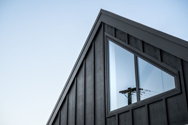 Close-up exterior view of a dark charcoal-clad gable end with vertical board-and-batten-style cladding, a large rectangular window, and a sharply pitched roofline against a clear sky.