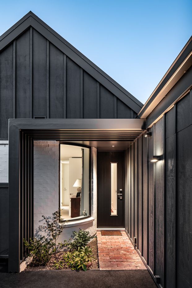 A narrow covered entrance passage between dark-clad building volumes, featuring steep gabled roofs, vertical charcoal cladding, a black-framed glazed opening, recessed entry door, brick paving, and a small planted bed. The contemporary exterior is photographed against a clear blue sky.