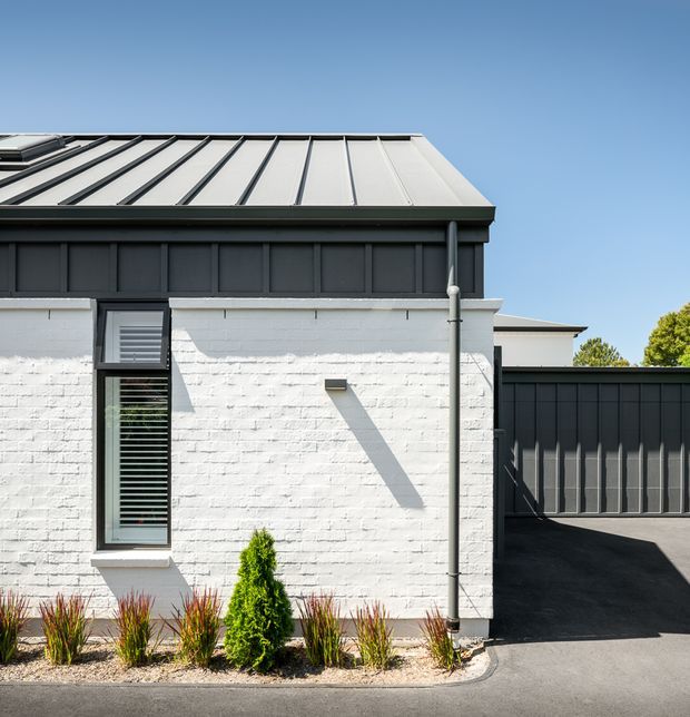 A close exterior view of a renovated single-storey building with white-painted brick walls, dark vertical-panel cladding, a standing-seam metal roof, narrow louvred window, exposed downpipe, and a dark driveway or paved access area. Sparse ornamental planting lines the base under a clear blue sky.