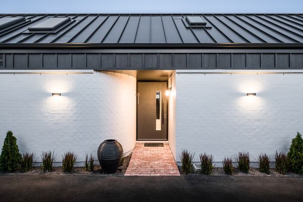 Front exterior view of a low, contemporary building with a dark standing-seam metal roof and matching fascia, white painted brick walls, and a recessed entry door reached by a red brick path. Minimal wall-mounted lighting, dark paving, a large ceramic urn, and restrained planting frame the entrance.