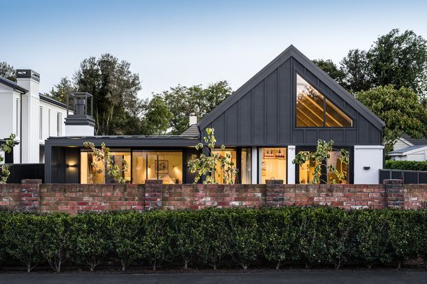 Street-facing exterior photograph of a contemporary single-storey house with a prominent steep gabled volume clad in dark vertical panels, warm-lit glazing, and a lower connecting wing. A low red brick boundary wall and manicured hedge frame the frontage, with neighbouring homes and mature trees in the background.