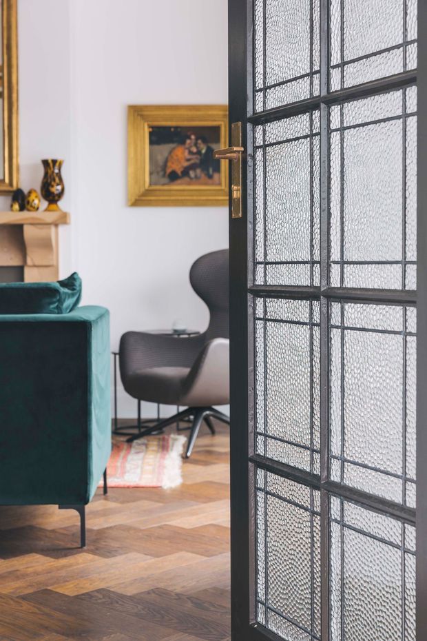 A close interior view through a dark-framed glazed door into a refined living room. The room features a deep teal upholstered sofa, a sculptural dark lounge chair, a timber herringbone parquet floor, white walls, a traditional mantel with decorative objects, and a framed artwork. The door has black metal framing, textured patterned glass, and brass hardware, creating a strong architectural foreground element.