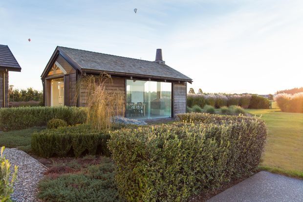 A compact single-storey garden pavilion or small house set within a formal landscaped garden. The building has dark timber weatherboard cladding, a pitched gabled roof with dark shingles or tiles, a chimney, and large glazed openings. Clipped hedges, gravel paths, ornamental planting, and a lawn surround the structure, with warm low-angle sunlight creating a tranquil rural garden setting.