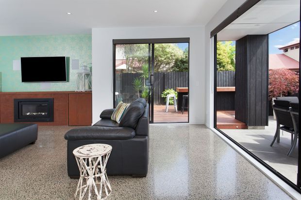 A bright contemporary living room with polished terrazzo-style flooring, a black leather sofa and armchair, a timber built-in media wall with fireplace, and a large corner of sliding or folding glazing opening to a timber deck and outdoor dining area. A turquoise textured feature wall adds colour, while dark vertical timber screening frames the exterior.