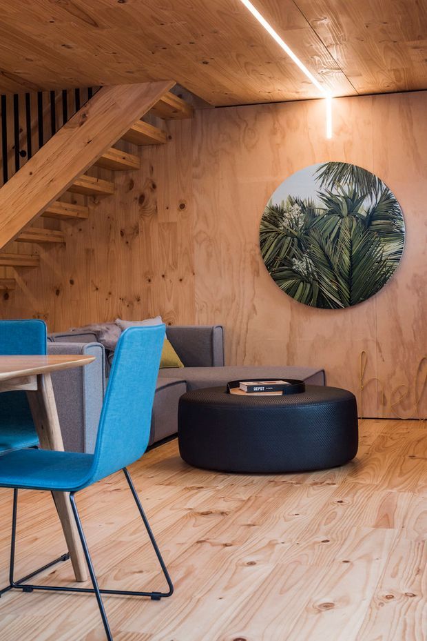 A compact, warmly lit lounge and dining area lined throughout with natural plywood. A timber staircase with open treads rises along the left wall beneath a plywood ceiling. The room contains a grey sofa, vivid blue dining chairs, a light timber table, and a low dark upholstered ottoman. A round wall artwork or mirror depicting foliage adds a focal point, while a linear ceiling light provides contemporary illumination.