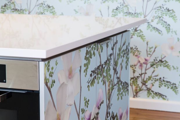 Close-up of a contemporary built-in cabinet or kitchen island with a pale benchtop, dark trim, and a decorative botanical floral panel on the side. A black integrated appliance or drawer front is partially visible, with timber flooring and matching botanical wall imagery in the background.