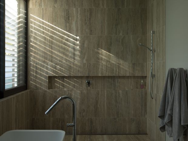 A refined contemporary bathroom featuring large-format stone-look wall and floor tiles in warm taupe-brown tones. The scene includes a partially visible freestanding bathtub with a chrome floor-mounted tap, a recessed wall niche, and a wall-mounted shower with handheld hose. A dark-framed louvred window on the left casts strong linear sunlight across the tiled surfaces, while grey towels hang on the right.