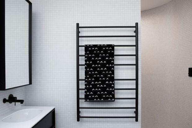A contemporary bathroom featuring white small-format tiled walls, a wall-mounted black heated towel rail with a black patterned towel, a white vanity basin with dark cabinetry, and a large black-framed mirror. The room has a clean monochrome palette and minimal fixtures.