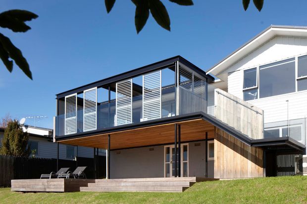Exterior view of a contemporary multi-level residential extension with a cantilevered upper volume, extensive glazing, horizontal louvres, dark metal framing, and warm timber soffits and cladding. The lower level opens to a lawn and timber deck, while the neighbouring white weatherboard house is visible to the right.