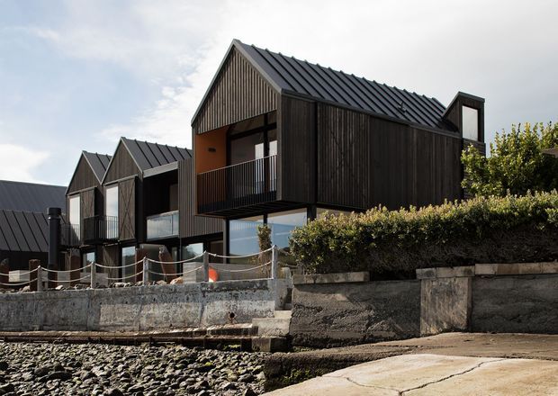 Exterior view of a contemporary multi-unit residential development on a rocky waterfront edge. The buildings feature repeated steep gabled forms, dark vertical timber cladding, dark metal pitched roofs, large glazed openings, and elevated balconies. Concrete retaining walls, a paved access area, and shoreline rocks are visible in the foreground.