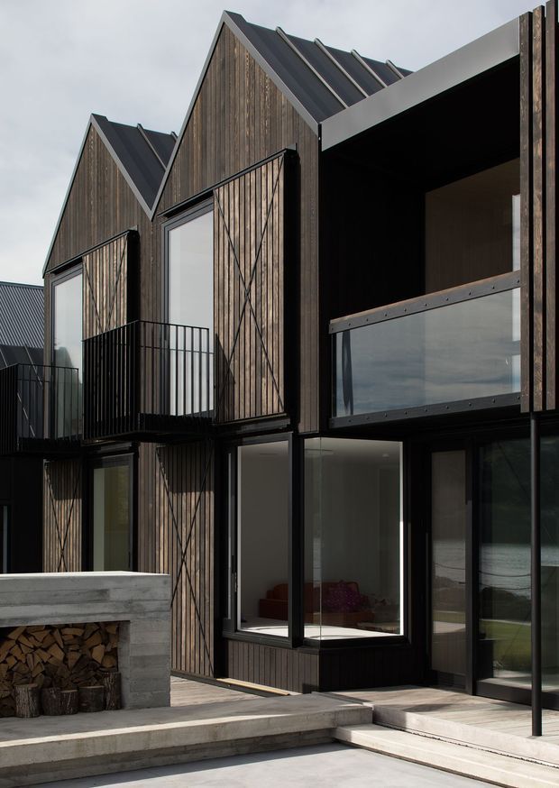 Exterior view of a contemporary multi-level residential building composed of several steep gabled volumes. The facade combines dark vertical timber cladding with dark metal roofing, black-framed glazing, projecting balconies, and a glazed upper-level terrace. A substantial concrete outdoor fireplace and paved terrace occupy the foreground. The architecture has a strong geometric, clustered form and appears to sit beside a body of water visible through the glazing.