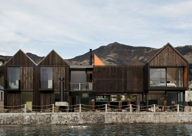 A contemporary multi-unit waterfront building with a striking series of steep gabled roof forms and dark vertical timber screening. The facade combines timber-clad upper volumes with dark-framed glazing and recessed balconies at ground level. A rope-railed waterfront promenade and retaining wall sit directly in front, with rugged mountains visible behind the building.