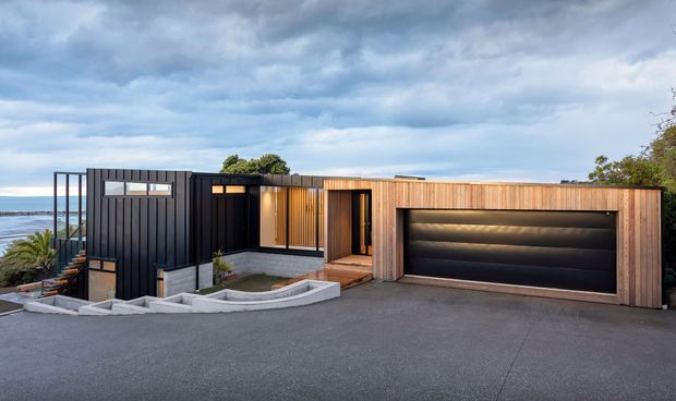 Exterior view of a low-profile contemporary house on a sloping coastal site, with dark vertical cladding, warm timber-clad sections, large glazed openings, a broad black double garage door, and a paved driveway. The ocean is visible beyond the left side of the house.