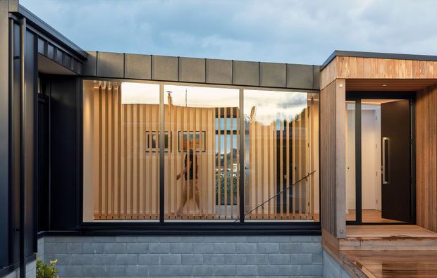 Exterior view of a contemporary residential entry and circulation zone. The composition features dark metal-framed glazing, vertical timber screening, a timber-clad entry volume with a recessed black door, a low grey masonry base, and a flat roofline. Interior stairs and a glimpse of distant landscape are visible through the glazing.