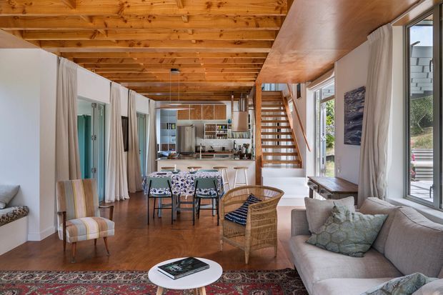 Open-plan residential interior combining a living room, dining area and kitchen beneath a warm timber-lined ceiling. The room features polished timber flooring, white walls, large windows with sheer curtains, a staircase with timber treads, a patterned rug, upholstered seating, a woven armchair and a compact kitchen with an island bench.