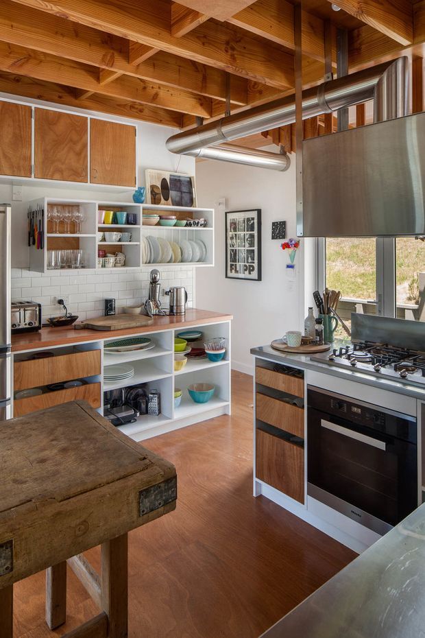 A warm, eclectic kitchen with exposed timber ceiling beams, polished metal ductwork, white subway-tile backsplash, open white shelving filled with dishes and glassware, timber cabinetry, and stainless-steel appliances. The compact layout includes a freestanding worktable in the foreground, a gas cooktop and oven, and a timber-toned floor. The space combines workshop-like industrial details with rustic and mid-century-inspired furnishings.