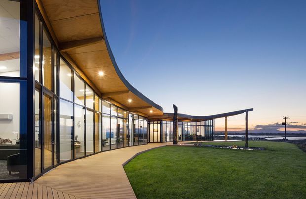 A contemporary curved building exterior at dusk, featuring extensive floor-to-ceiling glazing with dark slender frames, a warm timber soffit and deck walkway, and a broad manicured lawn leading toward a waterfront or body of water. A simple open pergola-like structure extends across the garden, while interior furnishings are visible through the glass.