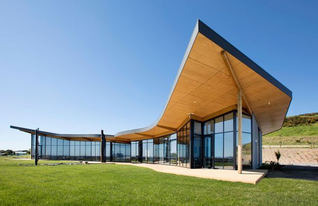A contemporary low-rise building set in a landscaped grassy site, featuring expansive dark-framed floor-to-ceiling glazing and a dramatic sculptural roof with broad timber-lined soffits. The roof curves and projects outward to form covered outdoor areas, while exposed slender supports and connected glazed wings create a strong indoor-outdoor relationship. A sloping planted embankment is visible behind the building under a clear blue sky.