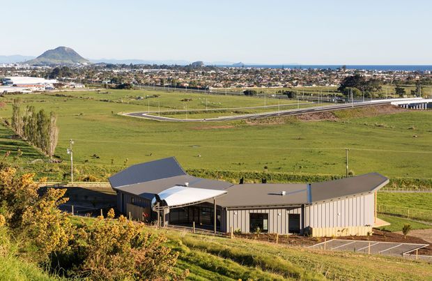 Aerial view of a low-rise contemporary building with dark and neutral metal-clad wings, a prominent curved white canopy, and surrounding paved access areas. The building sits on an expansive rural or semi-rural site, with open green fields, a nearby rail or road corridor, a developed settlement, coastline or large body of water, and a prominent hill visible in the distance.
