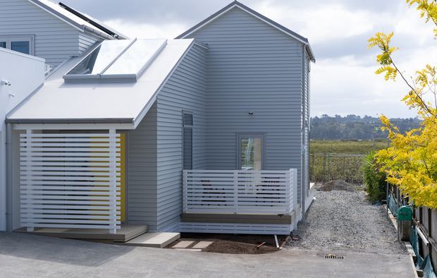 Exterior view of a compact contemporary residential dwelling with pale grey horizontal weatherboard cladding, steep pitched gable roofs, a projecting covered entry or carport-like structure, and a small raised balcony enclosed by horizontal slatted screening. The home sits beside a gravel side path and landscaped boundary, with rural or semi-rural vegetation visible beyond.