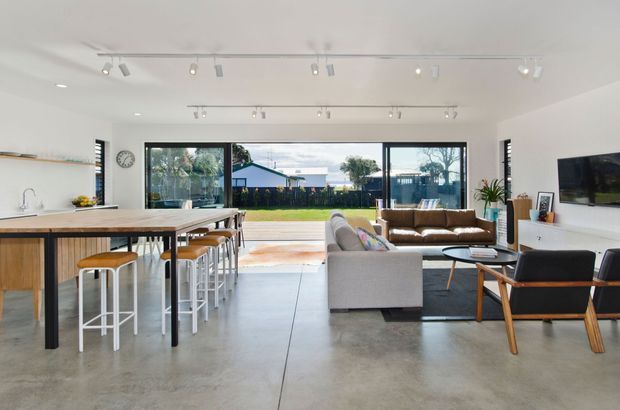 Bright open-plan contemporary living space combining a kitchen, dining island and lounge. The room has polished concrete flooring, white walls and ceiling, extensive black-framed glazing opening to a lawn, a timber kitchen island with white benchtop, bar stools, a grey sofa, brown leather sofa, and mid-century-inspired lounge chairs. Track lighting runs across the ceiling, with a wall-mounted television and minimal built-in storage.