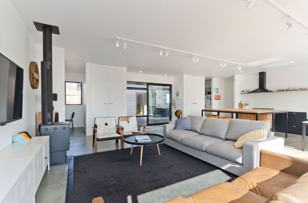 Bright open-plan contemporary living space with a large grey sectional sofa, black area rug, timber lounge chairs, wall-mounted television, and a compact dining and kitchen area beyond. White built-in cabinetry, track lighting, a black freestanding wood heater, dark-framed glazing, and a black kitchen rangehood create a clean modern interior.