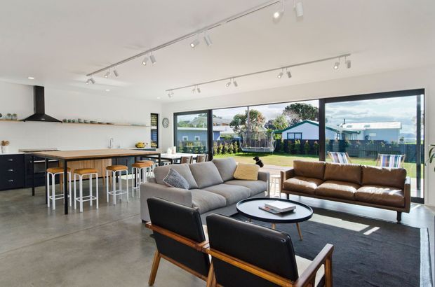 Bright open-plan contemporary living space combining a kitchen, dining island and living room. The room has polished concrete flooring, white walls and ceiling, black-framed full-height glazing, a timber kitchen island with bar stools, a large grey sofa, brown leather sofa and mid-century-style lounge chairs. Track lighting runs across the ceiling, and the glazing opens views to a landscaped suburban backyard.