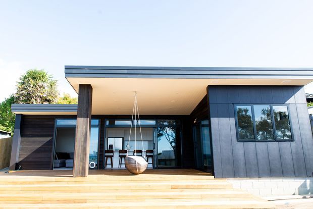 Front exterior view of a contemporary single-storey home with a flat roof, dark vertical cladding, extensive glazing, and a broad timber deck with steps. A covered outdoor living area includes a hanging chair, while the interior dining area is visible through the glazed doors.