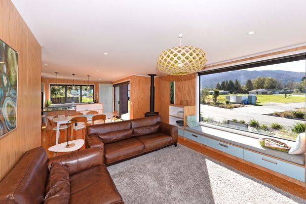 A bright mid-century-inspired open-plan living and dining area with extensive warm timber wall panelling, a large picture window overlooking a mountainous suburban landscape, and built-in low cabinetry beneath the glazing. The room features brown leather sectional seating, a round dining table with wooden chairs, a sculptural woven pendant light, patterned artwork, and a grey area rug.