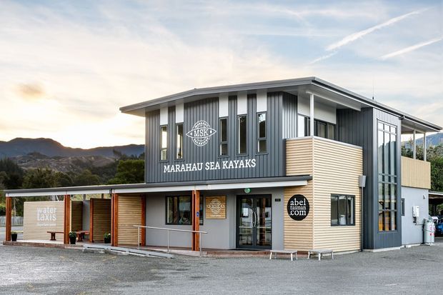Exterior view of a compact two-storey commercial building housing Marahau Sea Kayaks. The building has dark grey vertical corrugated cladding, light timber-toned horizontal wall panels, extensive windows, a shallow low-pitched roof with broad eaves, covered ground-floor verandah or loading area, and prominent business signage. A mountainous landscape is visible in the background.