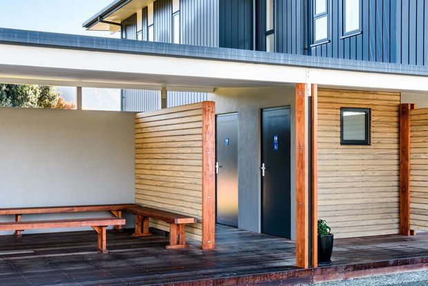 Exterior view of a contemporary multi-level building entrance with a covered timber deck and sheltered seating area. The facade combines dark blue-grey vertical metal cladding, horizontal light timber wall cladding, exposed warm timber posts, and dark decking. Two dark entry doors and a small window are visible beneath the broad flat canopy.