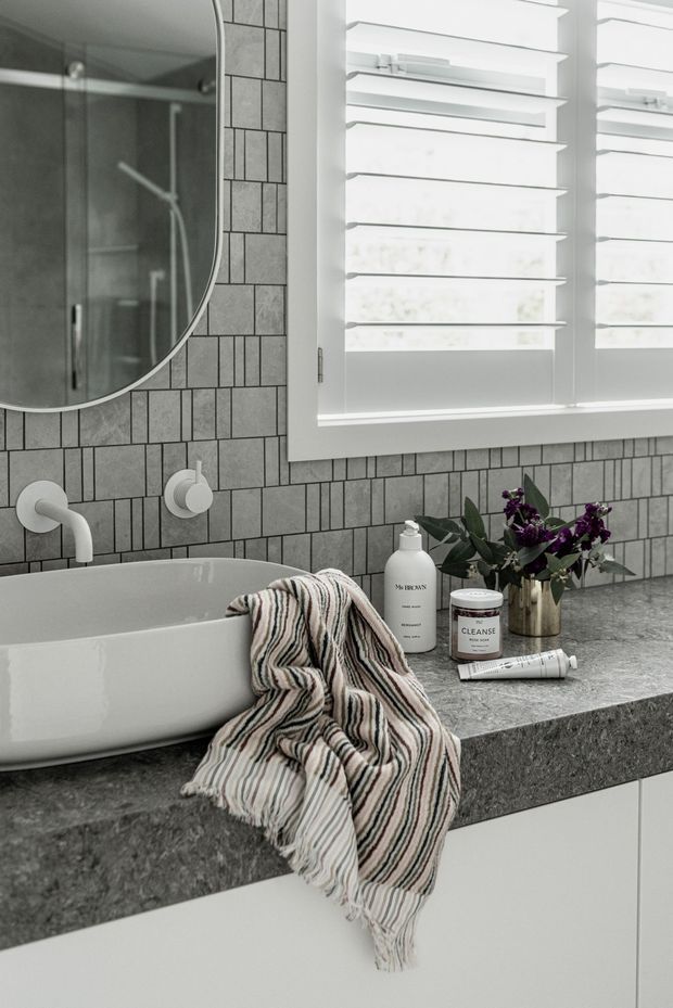 Close-up view of a refined bathroom vanity featuring a white vessel basin, wall-mounted white tapware, grey rectangular mosaic tile splashback, dark grey stone benchtop, white cabinetry, and a large rounded mirror. White plantation shutters provide filtered natural light, while a striped towel, toiletries, and a small floral arrangement add softness.