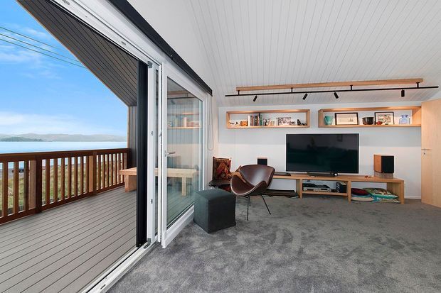 Bright modern living room opening onto a timber-decked balcony through wide sliding glass doors. The room features grey carpet, white walls and ceiling, a long timber media console with wall-mounted television, recessed display shelving, a leather lounge chair, and a linear track light. Mountainous scenery is visible beyond the balcony.