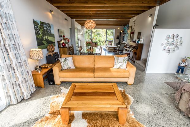A warm open-plan living room with a tan leather sofa, substantial timber coffee table, patterned cushions, and a cowhide rug on a polished speckled floor. The room has exposed timber ceiling beams, white walls, extensive rear glazing overlooking greenery, a dining area beyond the sofa, and eclectic decorative furnishings.