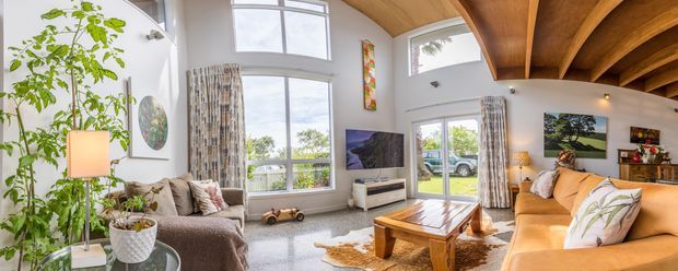 Bright, spacious living room with double-height glazing, a warm timber-beamed ceiling, large sectional seating, television console, coffee table, and indoor plants. The room opens toward an exterior garden or patio through glazed doors and has a relaxed, warm contemporary feel.