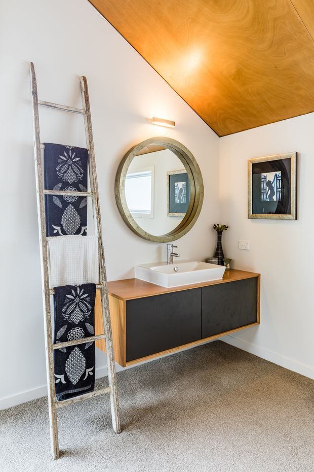 Bright bathroom featuring a floating timber vanity with dark flat-front cabinetry, a white vessel basin, chrome tapware, and a large round framed mirror. White walls, a warm timber-clad sloping ceiling, carpeted floor, wall artwork, and a rustic ladder used as a towel rack create a warm contemporary interior.