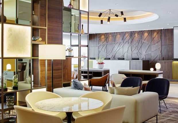 A refined contemporary lounge and dining area with a round marble-topped dining table, upholstered dining chairs, sofas, lounge chairs, and a dark timber and glass open shelving partition. The room features a dramatic textured feature wall, sculptural linear pendant lighting, layered recessed ceiling details, and warm neutral finishes.