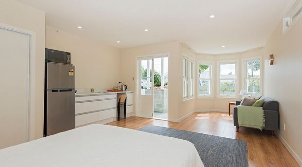 Bright, open-plan studio-style interior combining a compact white kitchen with stainless-steel refrigerator, a sleeping area, and a small living zone. The room features warm timber flooring, pale walls, recessed ceiling lights, large bay-style windows, and a glazed door opening to an exterior balcony or terrace. A grey sofa with a green throw sits beside the windows, while the bed occupies the foreground.
