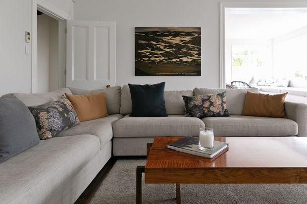 A bright, refined living room featuring a large light-grey L-shaped sectional sofa with neutral and patterned cushions, a substantial dark timber coffee table, and a textured grey rug. White walls, traditional panelled doors and trim, and a dark landscape artwork create a calm, contemporary-traditional setting. A bright adjoining sunroom or glazed sitting area is visible beyond the living room.