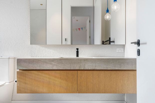 Bright contemporary bathroom vanity with a long floating timber cabinet, thick pale concrete benchtop, black wall-mounted tap, and full-width mirrored storage above. Small white hexagonal mosaic tiles cover the wall, while globe pendant lights hang beside the mirror. A toilet is partially visible at left and a doorway opens behind the vanity.