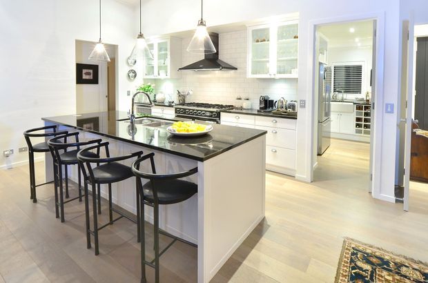 Bright, spacious white kitchen with a large central island topped in dark stone and paired with black bar stools. The room features white shaker-style cabinetry, a white subway-tile splashback, glass-front upper cabinets, stainless steel appliances, pendant lighting, and pale timber flooring. A pantry or utility space is visible through the doorway, with a glimpse of an adjacent room beyond.